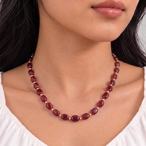 May include: A necklace featuring a series of smooth, oval-shaped deep red gemstones alternating with small, polished gold-toned spherical beads. The necklace rests against the neck, showcasing a consistent pattern of red stones and gold accents.