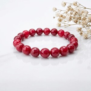 Könnte beinhalten: Ein kreisförmiges Armband, das aus zahlreichen runden, facettierten Perlen in einer tiefen, leuchtenden roten Farbe besteht. Die Perlen sind zu einer durchgehenden Schlaufe aufgereiht und weisen eine polierte, reflektierende Oberfläche auf.