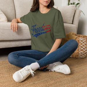 May include: An olive green short-sleeve t-shirt featuring the word Freedom printed in a distressed blue and red font with a star pattern integrated into the lettering. The shirt is paired with dark blue denim jeans and white lace-up trainers.