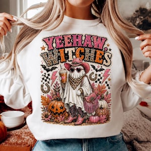 May include: A white sweatshirt featuring a graphic print with the text YEEHAW WITCHES. The design shows a ghost wearing a pink cowboy hat, heart-shaped sunglasses, and a leopard print bandana, holding a drink, with pumpkins, stars, and bats.