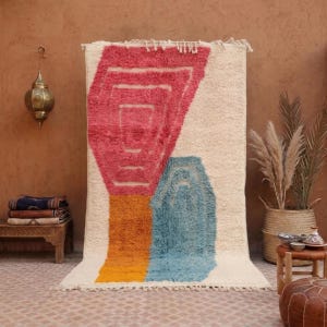 Könnte beinhalten: Ein rechteckiger Teppich mit cremefarbenem Hintergrund und einem großen, abstrakten geometrischen Design in Rosa-, Orange- und Blautönen. Der Teppich hat eine dicke, hochflorige Textur und ist an der Oberkante mit Fransen versehen.
