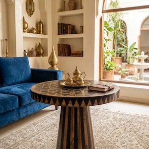 May include: A round wooden table with a dark and light striped pedestal base and a geometric patterned top. On the table sits a gold-colored metal tea set on a tray and a small closed book. The table is positioned in a room with a blue sofa.