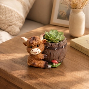 Op de afbeelding: Een bruin hooglandrund-figuurtje bevestigd aan een bruine tonvormige plantenbak. De plantenbak bevat een groene vetplant en heeft een kleine rode paddenstoel aan de basis. De ton heeft een donkerbruine band met kleine zilverkleurige accenten.