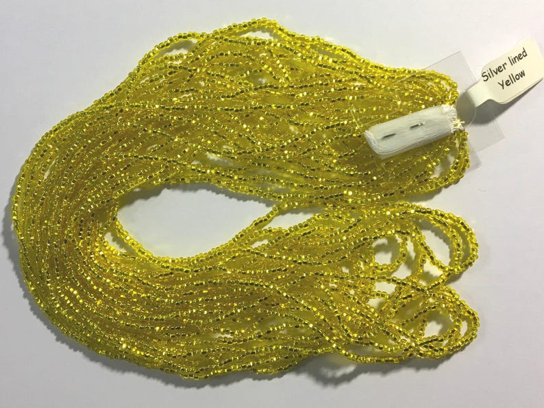 Puede incluir: Un gran carrete de cuentas de semillas de color amarillo brillante con un borde plateado. Las cuentas est&aacute;n dispuestas en una pila suelta, con algunas hebras superpuestas. La etiqueta dice "Silver lined Yellow".