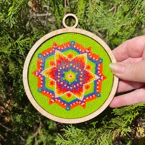K&ouml;nnte beinhalten: Ein runder Holzanh&auml;nger mit gr&uuml;nem Hintergrund und einem bunten Perlenmandala-Design. Das Mandala zeigt ein Sternmuster mit roten, orangefarbenen, gelben, blauen und violetten Perlen.