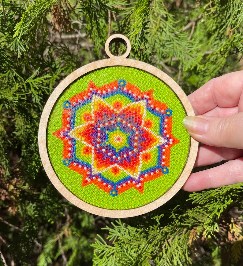 K&ouml;nnte beinhalten: Ein runder Holzanh&auml;nger mit gr&uuml;nem Hintergrund und einem bunten Perlenmandala-Design. Das Mandala zeigt ein Sternmuster mit roten, orangefarbenen, gelben, blauen und violetten Perlen.