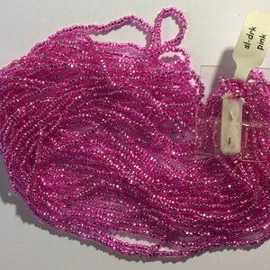 Puede incluir: Un mont&oacute;n grande de cuentas de semillas rosas con un brillo met&aacute;lico. Las cuentas est&aacute;n dispuestas en un mont&oacute;n suelto y fluido. Una peque&ntilde;a etiqueta blanca con el texto "sl-drk Pink" es visible en la esquina superior derecha de la imagen.