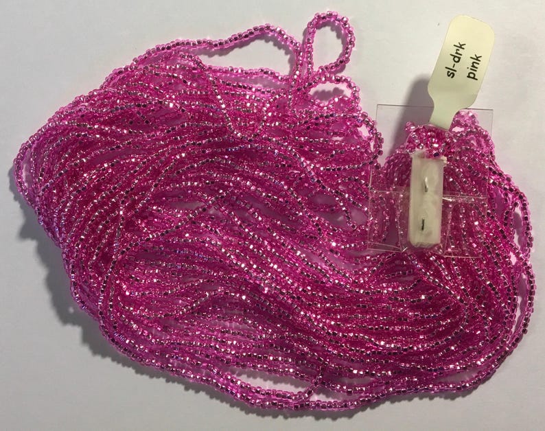 Puede incluir: Un mont&oacute;n grande de cuentas de semillas rosas con un brillo met&aacute;lico. Las cuentas est&aacute;n dispuestas en un mont&oacute;n suelto y fluido. Una peque&ntilde;a etiqueta blanca con el texto "sl-drk Pink" es visible en la esquina superior derecha de la imagen.