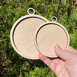 K&ouml;nnte beinhalten: Zwei unfertige runde Holzornamente mit einem Loch oben zum Aufh&auml;ngen. Das gr&ouml;&szlig;ere Ornament befindet sich hinter dem kleineren.