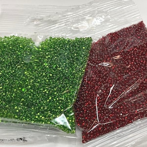 Puede incluir: Dos bolsas de cuentas de semillas, una verde y otra roja. Las bolsas son de pl&aacute;stico transparente y las cuentas son visibles.