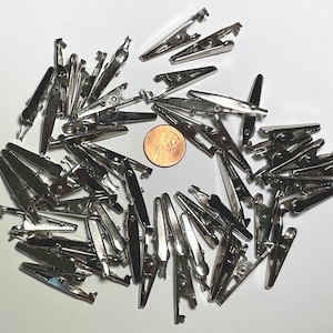 May include: A large group of silver-toned alligator clips, also known as bulldog clips, are scattered on a white surface. The clips are approximately 1 inch in length. A penny is included for scale.