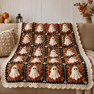Könnte beinhalten: Eine handgefertigte Häkeldecke mit einem Gitter aus quadratischen Motiven in schwarzem, orangefarbenem und grünem Garn. Jedes Quadrat enthält eine kleine, dreidimensionale weiße Geisterapplikation mit schwarzen Augen und einen cremefarbenen Rand.
