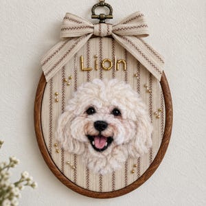 May include: An oval wooden embroidery hoop wall hanging featuring a needle-felted cream-coloured poodle portrait. The background fabric has vertical brown stripes and is adorned with small gold beads. The word LION is spelled out in gold beads.
