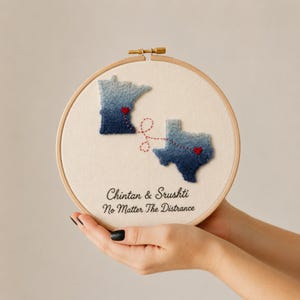 May include: A circular wooden embroidery hoop containing cream fabric with two blue ombre state shapes, Minnesota and Texas. Each state features a small red heart and a red stitched line connecting them. Text reads: Chintan & Srushti No Matter The Distrance.