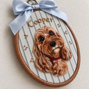 May include: An oval embroidery hoop frame featuring a needle-felted portrait of a brown dog with a red collar. The background fabric has a white and blue striped pattern. The name Cooper is spelled out in gold beads above the dog, with additional gold beads scattered around. A light blue satin ribbon is tied at the top.