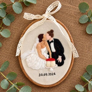 May include: An oval embroidery hoop featuring a needle-felted scene of a couple in wedding attire sharing a cake. The figure on the left wears a white dress and the figure on the right wears a black suit. The date 30.09.2024 is stitched below.