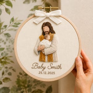 May include: A circular wooden embroidery hoop containing a fabric panel with a needle-felted figure of a person with long brown hair holding a swaddled infant. A white ribbon is tied at the top. The text Baby Smith and 25.12.2025 is embroidered below.