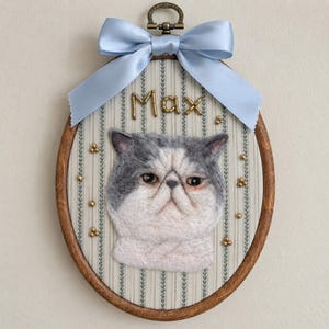 May include: An oval wooden embroidery hoop frame containing a needle-felted grey and white cat portrait on striped fabric. The name Max is spelled out in gold beads above the cat. A light blue satin ribbon bow is tied at the top metal hanger.