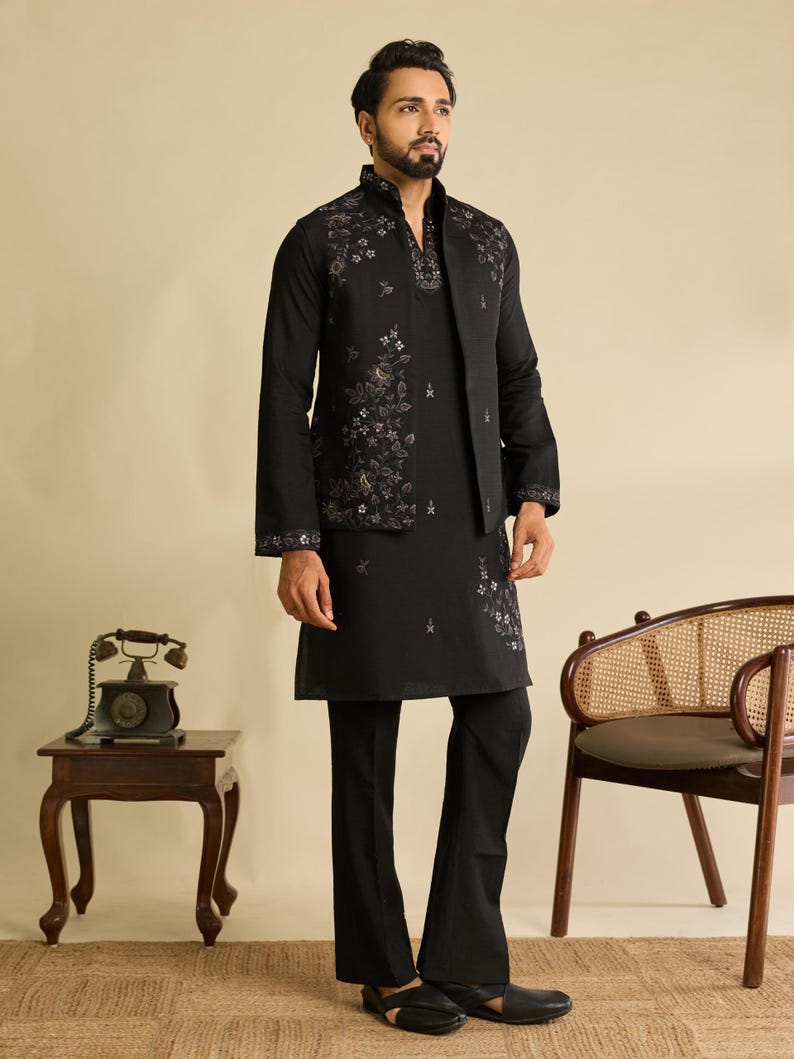 May include: A black long-sleeved kurta paired with matching black trousers and a sleeveless vest. The vest and kurta feature intricate floral embroidery in muted tones. The vest has a high collar, and the ensemble is completed with black sandals.