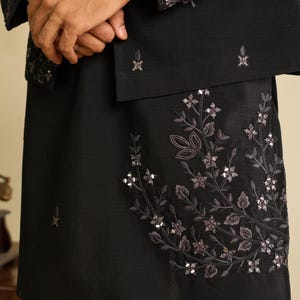 May include: A black long-sleeved garment featuring intricate floral embroidery and reflective silver-toned sequins. The design includes a detailed botanical pattern on the lower section and matching decorative embroidery on the sleeve cuffs.