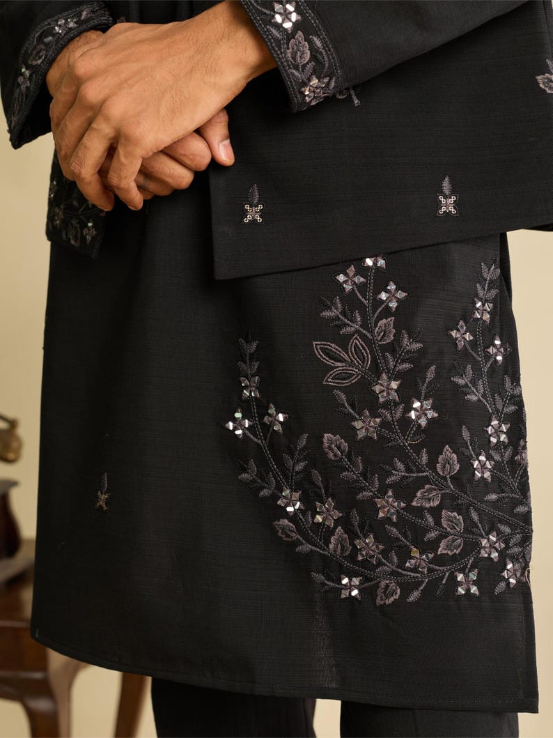 May include: A black long-sleeved garment featuring intricate floral embroidery and reflective silver-toned sequins. The design includes a detailed botanical pattern on the lower section and matching decorative embroidery on the sleeve cuffs.