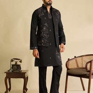May include: A black long-sleeved kurta paired with matching black trousers and a sleeveless vest. The vest and kurta feature intricate floral embroidery in muted tones. The vest has a high collar, and the ensemble is completed with black sandals.