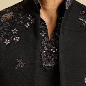 May include: A black long-sleeved garment featuring a high mandarin collar and a V-neck opening. The fabric is adorned with intricate dark floral embroidery and small, reflective metallic embellishments along the neckline and shoulders.