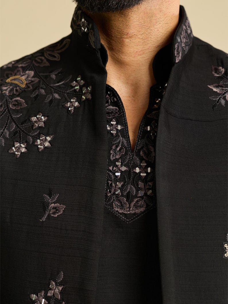 May include: A black long-sleeved garment featuring a high mandarin collar and a V-neck opening. The fabric is adorned with intricate dark floral embroidery and small, reflective metallic embellishments along the neckline and shoulders.