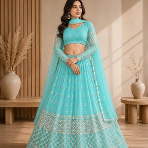 May include: A turquoise lehenga choli set featuring a long-sleeved blouse and a floor-length skirt, both adorned with intricate silver embroidery and mirror work. The ensemble includes a matching sheer dupatta draped over the shoulders.