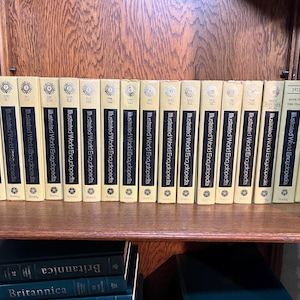 May include: A set of seventeen cream-colored Illustrated World Encyclopedia volumes arranged in a row, featuring black spine labels with gold text, numbered one through fifteen, plus two 1973 and 1974 Book of the Year editions by Bobley.