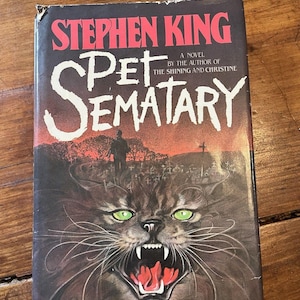 Könnte beinhalten: Ein Taschenbuch mit dem Titel Pet Sematary von Stephen King. Das Cover zeigt eine düstere Illustration einer knurrenden Katze mit leuchtend grünen Augen und scharfen Zähnen vor der Silhouette eines Friedhofs unter einem roten Himmel.