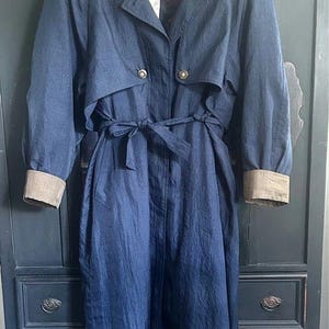 May include: A long, dark blue trench coat featuring a wide collar, a buttoned storm flap, and a tie-waist belt. The sleeves have contrasting tan cuffs, and the interior lining displays a dark red and black patterned design visible at the neck.