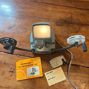 Peut inclure: Un éditeur de film 8mm Vernon 101 gris avec un écran éclairé, deux bobines de film et une manivelle. Sont inclus un livret d'instructions orange intitulé Vernon 8mm Movie Editor 101, un petit outil de montage en métal et une fiche Quik Splice.