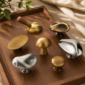 May include: A collection of seven cabinet knobs in various shapes and finishes, including brushed gold, polished silver, and a brass knob with a wooden handle. The set features organic, rounded, and mushroom-like forms arranged on a wooden surface.