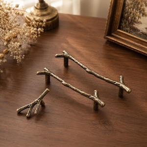 Peut inclure: Deux longues poignées de placard et un bouton plus petit, tous fabriqués en métal couleur laiton antique avec une finition texturée et organique imitant l'aspect de branches d'arbre naturelles. Chaque pièce présente une surface rugueuse et irrégulière.