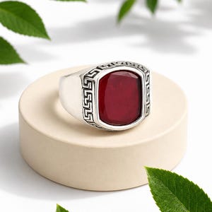 May include: A silver ring featuring a large, rectangular, faceted deep red stone set in the centre. The band is decorated with a repeating geometric Greek key pattern engraved into the metal surrounding the stone, creating a textured border.