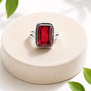 Puede incluir: Un anillo de plata con una gran piedra preciosa roja rectangular facetada engastada en un bisel de metal liso. La banda del anillo se divide en dos brazos delgados de plata pulida que se conectan al engaste central, creando un aspecto estructurado.