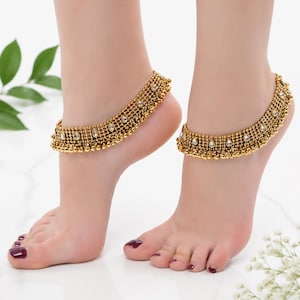 May include: A pair of gold-toned anklets featuring a multi-layered beaded chain design with small dangling metallic spheres and teardrop-shaped white stones set at regular intervals along the band, creating a textured and ornate appearance.