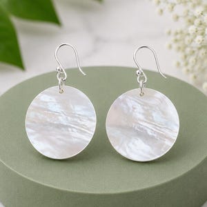 May include: A pair of drop earrings featuring circular, iridescent white mother-of-pearl discs suspended from silver-toned metal shepherd hook wires. The smooth, round shell components display a natural, swirling pearlescent texture.