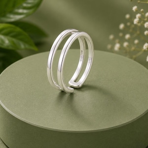 May include: A polished silver ring featuring a double-band design with an open gap. The metal surface is smooth and reflective, showcasing a clean, continuous loop structure. The interior of the band is clearly stamped with the hallmark 925.