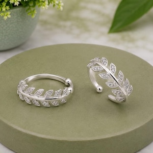Op de afbeelding: Twee zilverkleurige ringen met een delicaat bladvormig ontwerp. Elke ring is versierd met kleine, heldere, sprankelende steentjes die langs het oppervlak van de blaadjes zijn ingelegd, wat een doorlopend botanisch patroon vormt.