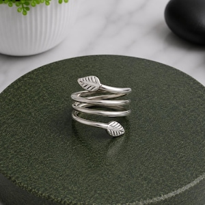 Può includere: Un anello color argento con un design a fascia a spirale che si avvolge attorno al dito. Ogni estremità dell'anello termina con un piccolo motivo a foglia dettagliato con venature incise, creando una struttura a spirale aperta continua.