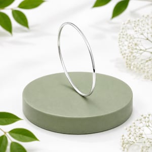 May include: A thin, polished silver-toned bangle bracelet with a smooth, rounded surface. The circular metal band is captured in a simple, minimalist design, showcasing a reflective metallic finish that catches the light along its entire circumference.