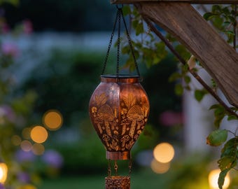 May include: A copper-coloured metal lantern shaped like a hot air balloon, featuring intricate cut-out designs of mushrooms and foliage. The lantern is suspended by thin black chains and emits a warm, glowing light from within its structure.