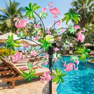 May include: A metal garden wind spinner featuring multiple pink flamingo figures and green palm leaf accents arranged on a circular frame. The structure is mounted on a black pole and includes a central light component with a textured glass cover.