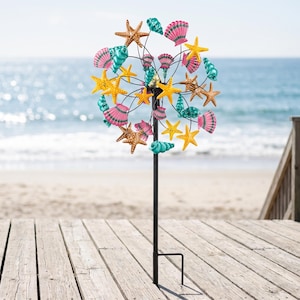 May include: A metal garden wind spinner featuring a circular arrangement of colorful starfish and seashell ornaments in shades of yellow, pink, and turquoise, mounted on a tall, slender black pole with a pronged base for ground installation.