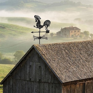 May include: A black metal weather vane featuring a rooster silhouette mounted on a directional arrow with cardinal points N, S, E, and W. The weather vane is positioned on the peak of a wooden barn roof against a backdrop of rolling hills.