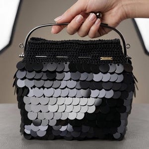 May include: A black crocheted handbag featuring a dense layer of overlapping circular black sequins. The bag has a metallic silver-toned arched handle and a small rectangular tag with the word Handmade attached to the upper crocheted section.