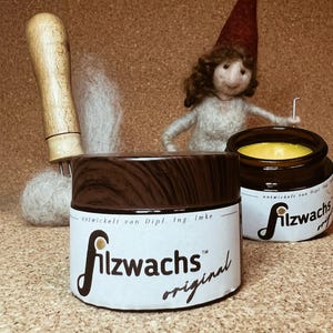Könnte beinhalten: Zwei Braunglastiegel mit dunkelbraunen Holzoptik-Deckeln, beschriftet mit Filzwachs original, entwickelt von Dipl. Ing. Imke. Ein Tiegel ist geöffnet und zeigt gelbes Wachs. Dahinter stehen ein Filzwerkzeug und eine kleine Filzfigur mit roter Mütze.