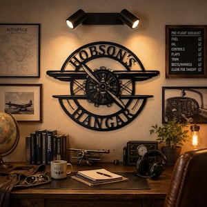 May include: A black metal wall sign featuring a circular design with an airplane propeller in the center and the text "HOBSON'S HANGAR" arched along the top and bottom. The sign is mounted on a wall above a desk with various aviation-themed items.